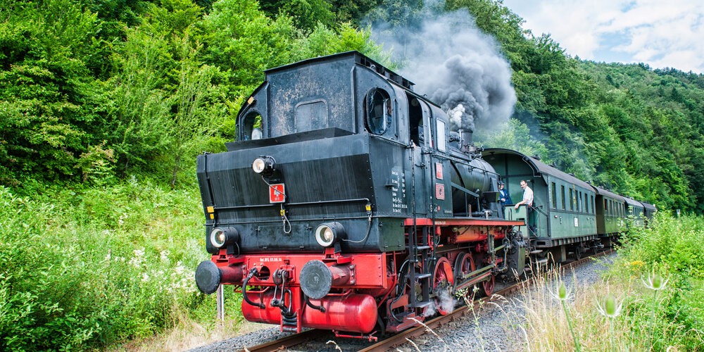 Eine alte schwarz-rote Dampfbahn stößt dunklen Rauch aus und fährt mit vier Waggons am Waldrand entlang.