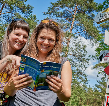 Zwei lachende Frauen stehen an einem Pfosten mit Wanderwegmarkierungszeichen und blättern in einem Reiseführer.