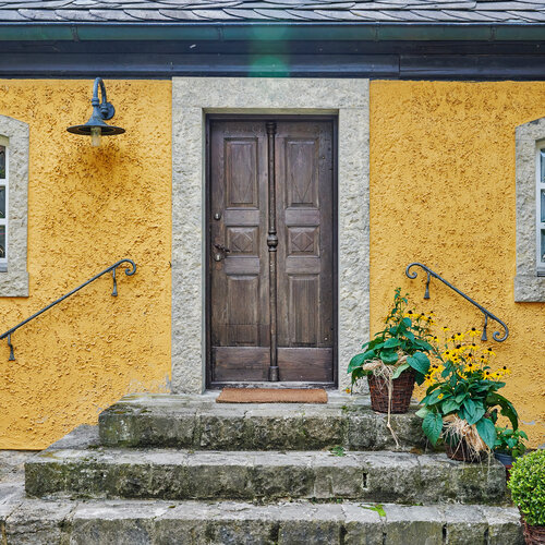 Eine alte hölzerne Eingangstür führt über Steintreppen in ein gelbes Haus.